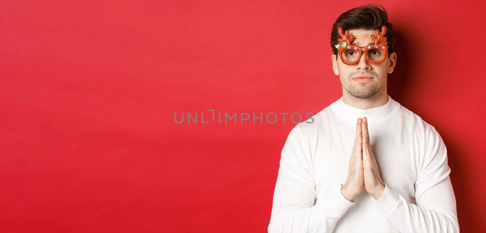 Close-up of cute guy in christmas party glasses, begging for help, need a favour, standing over red background.