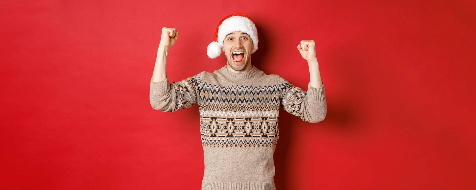 Image of cheerful handsome man in swearer and santa hat, celebrating new year, triumphing or winning something, raising hands up and shouting for joy, standing over red background by Benzoix