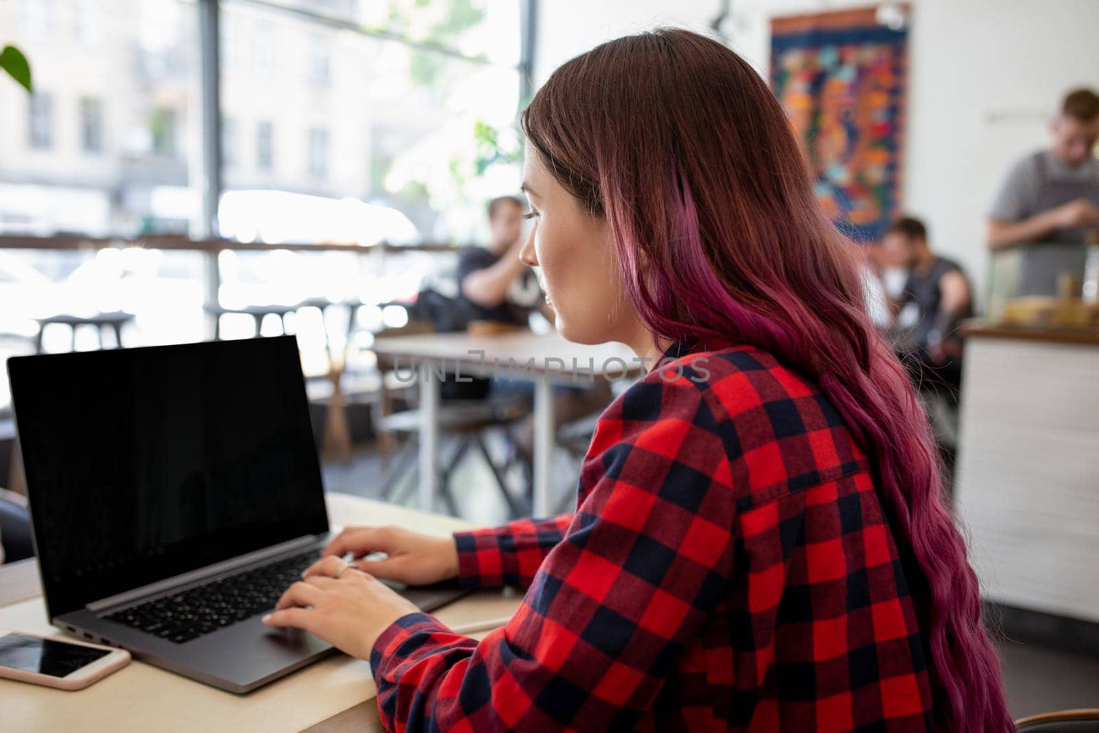 Back view of a young pink hair woman keyboarding on laptop computer with blank copy space screen while sitting in cafe. by nazarovsergey