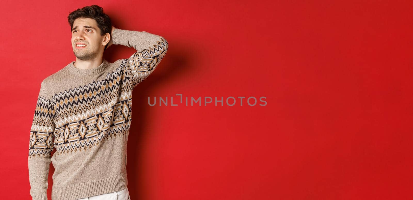 Image of puzzled and indecisive man in christmas sweater, looking at upper left corner, thinking about new year holidays gifts, standing over red background.