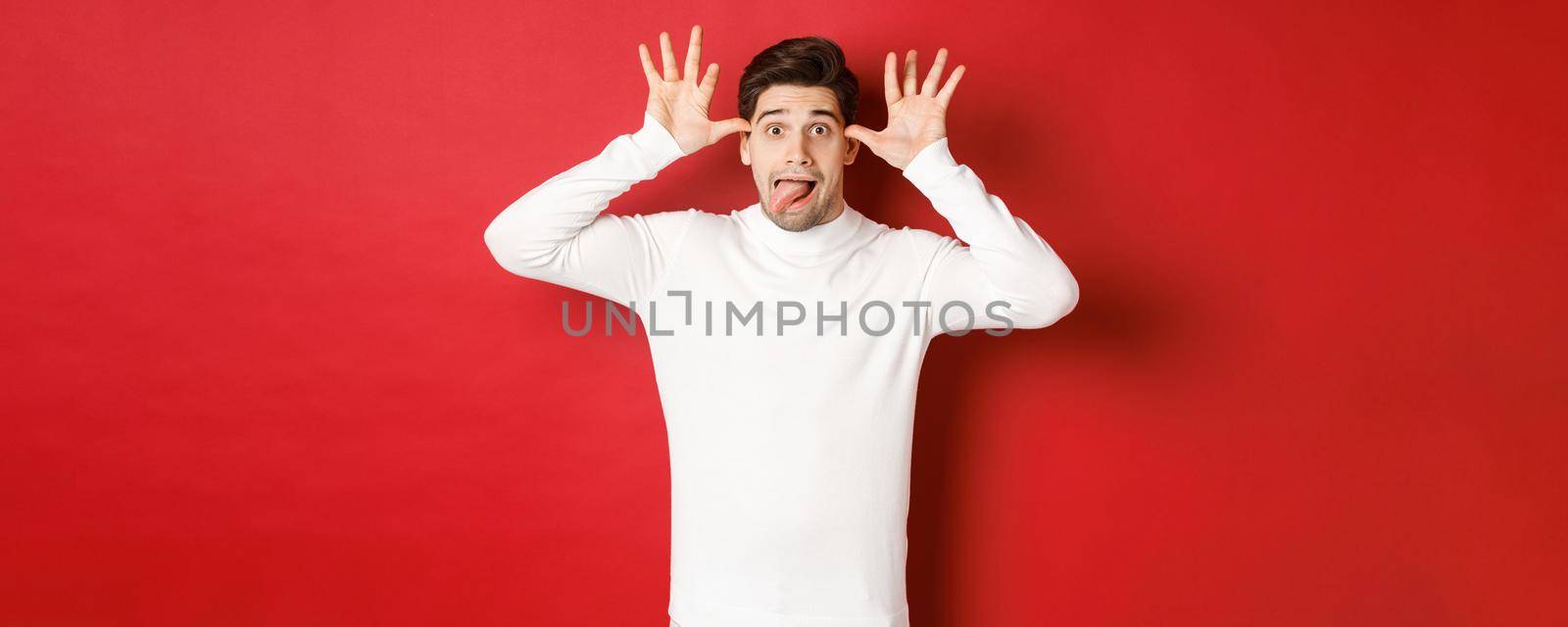 Portrait of funny caucasian guy, showing tongue and making faces, wearing white sweater, standing against red background by Benzoix
