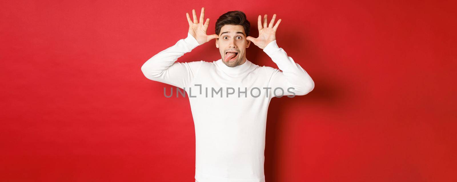 Portrait of funny caucasian guy, showing tongue and making faces, wearing white sweater, standing against red background.