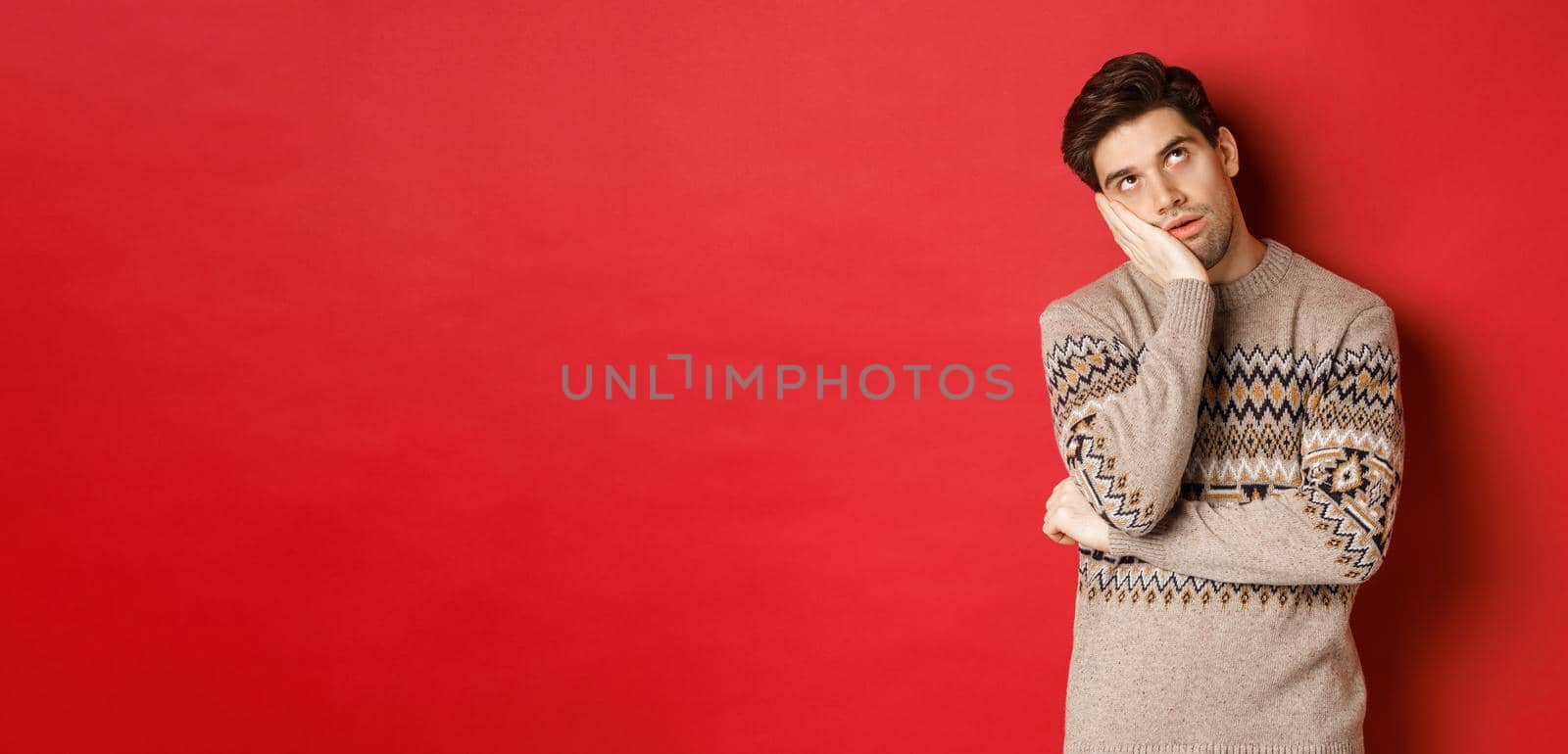 Image of annoyed young man in christmas sweater, roll eyes and leaning on hand, express displeasure and irritation, standing over red background.