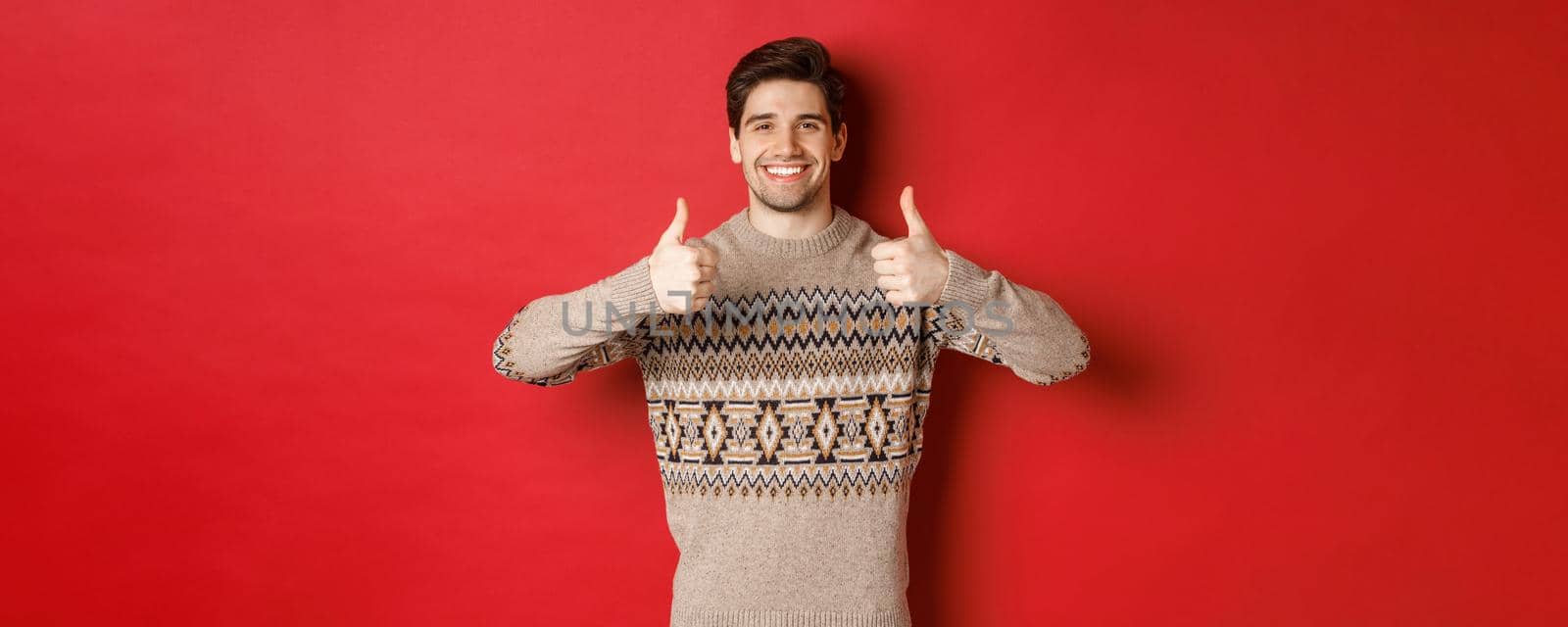 Portrait of happy and pleased handsome guy in christmas sweater, showing thumbs-up and nod in approval, smiling satisfied, standing over red background.