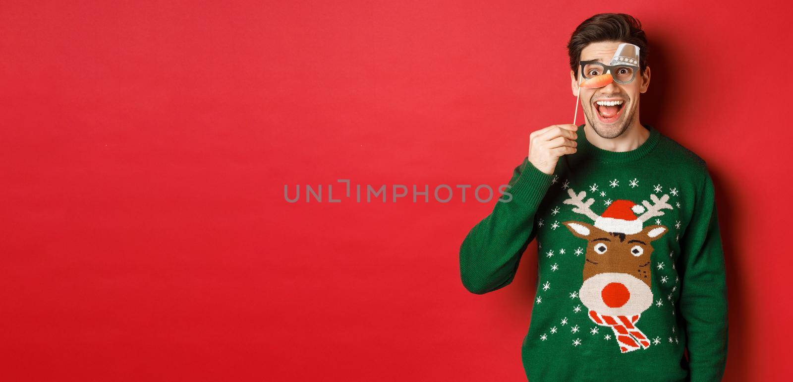 Portrait of amused handsome man in christmas sweater, holding funny party mask and smiling, celebrating winter holidays, standing over red background.