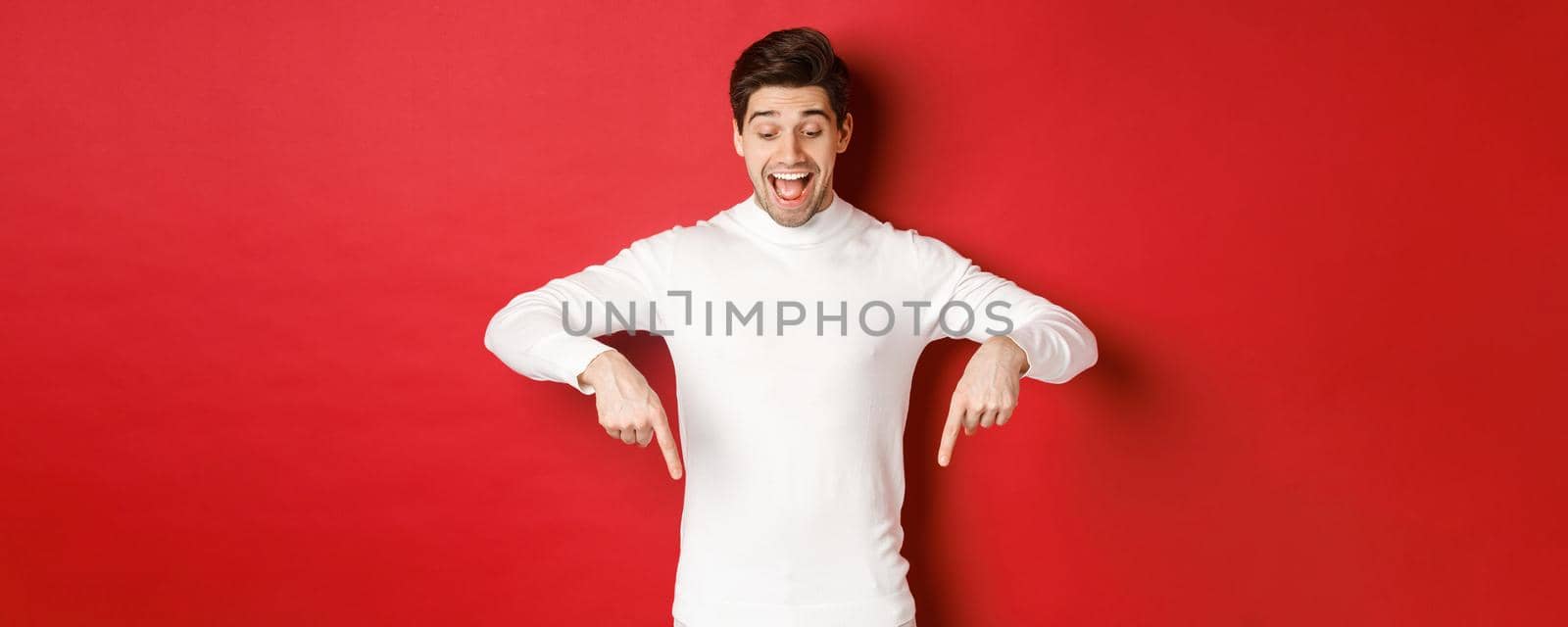 Portrait of happy good-looking man, pointing fingers down and looking at advertisement with excited smile, standing over red background.