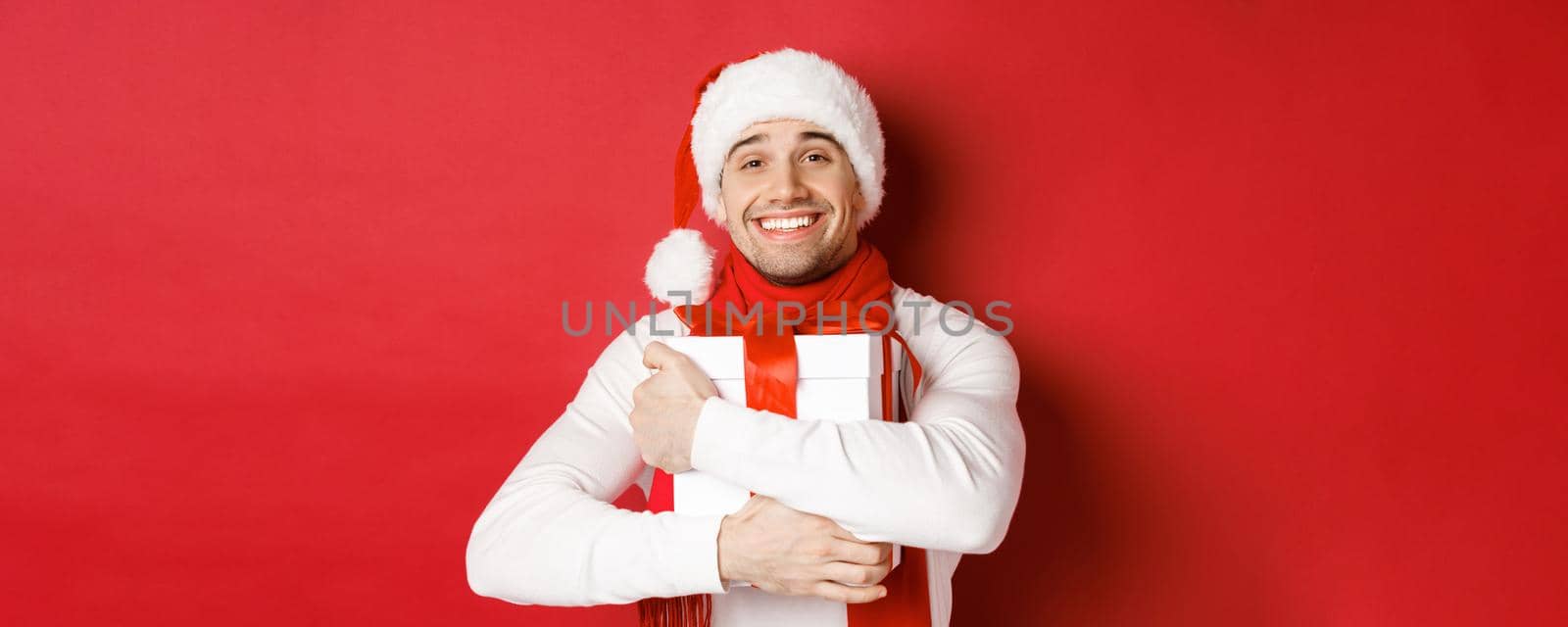 Concept of winter holidays, christmas and lifestyle. Image of lovely guy in santa hat and scarf, hugging his new year present and smiling flattered, standing over red background by Benzoix