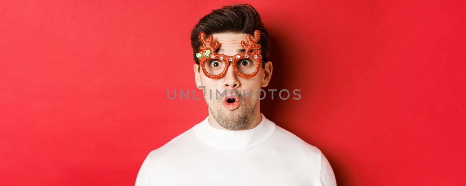 Concept of winter holidays, christmas and celebration. Close-up of attractive guy with bristle, wearing new year party glasses and looking impressed at promo offer, standing over red background by Benzoix