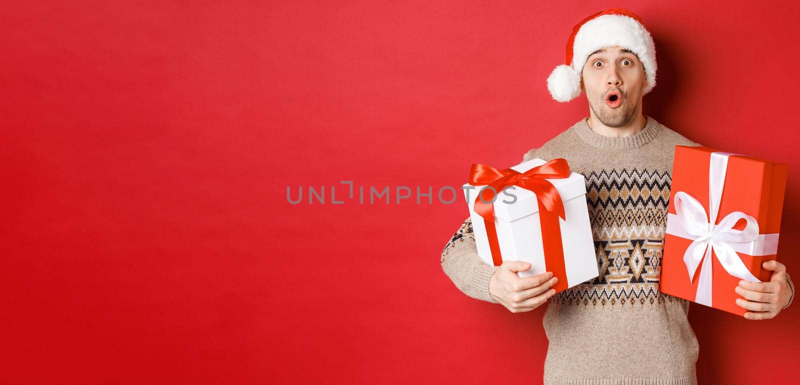 Concept of winter holidays, new year and celebration. Image of surprised attractive guy in santa hat and christmas sweater, receiving gifts, holding presents and looking amazed.