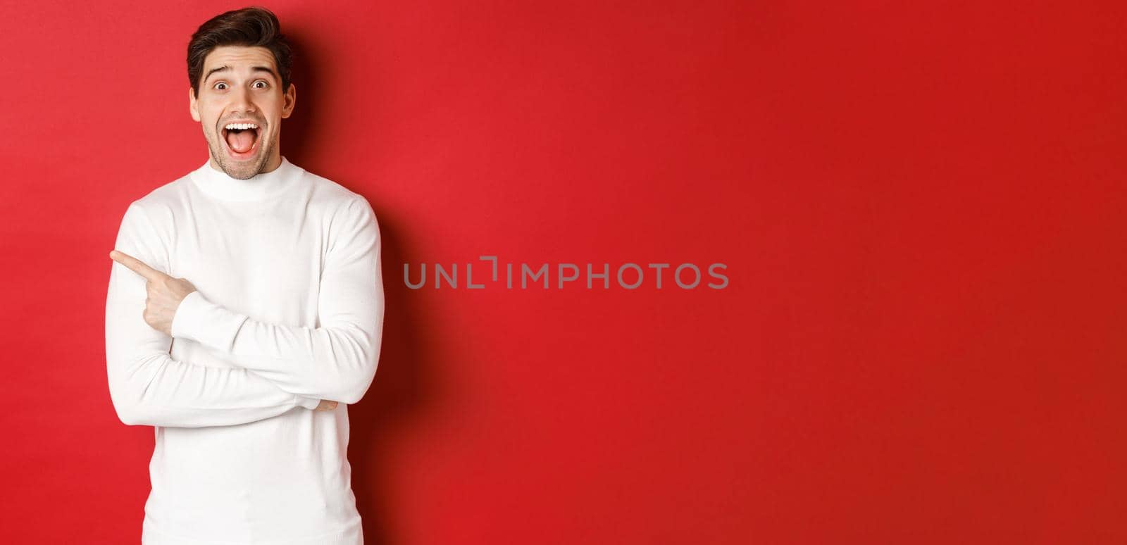 Concept of winter holidays, christmas and lifestyle. Amazed handsome man in white sweater showing logo, pointing finger left and looking excited, standing against red background.