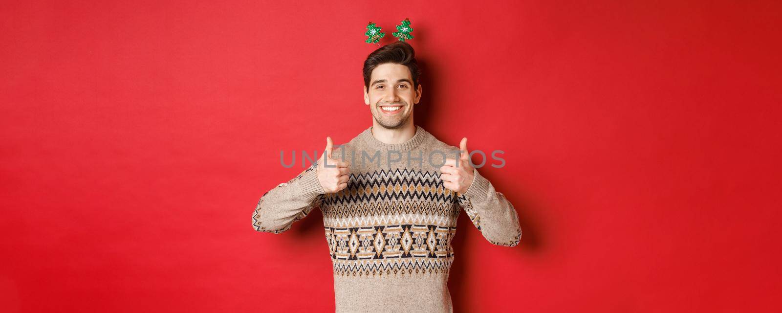 Concept of winter holidays, christmas and celebration. Cheerful bearded guy in sweater, showing thumbs-up in approval and smiling, enjoying new year party, red background.