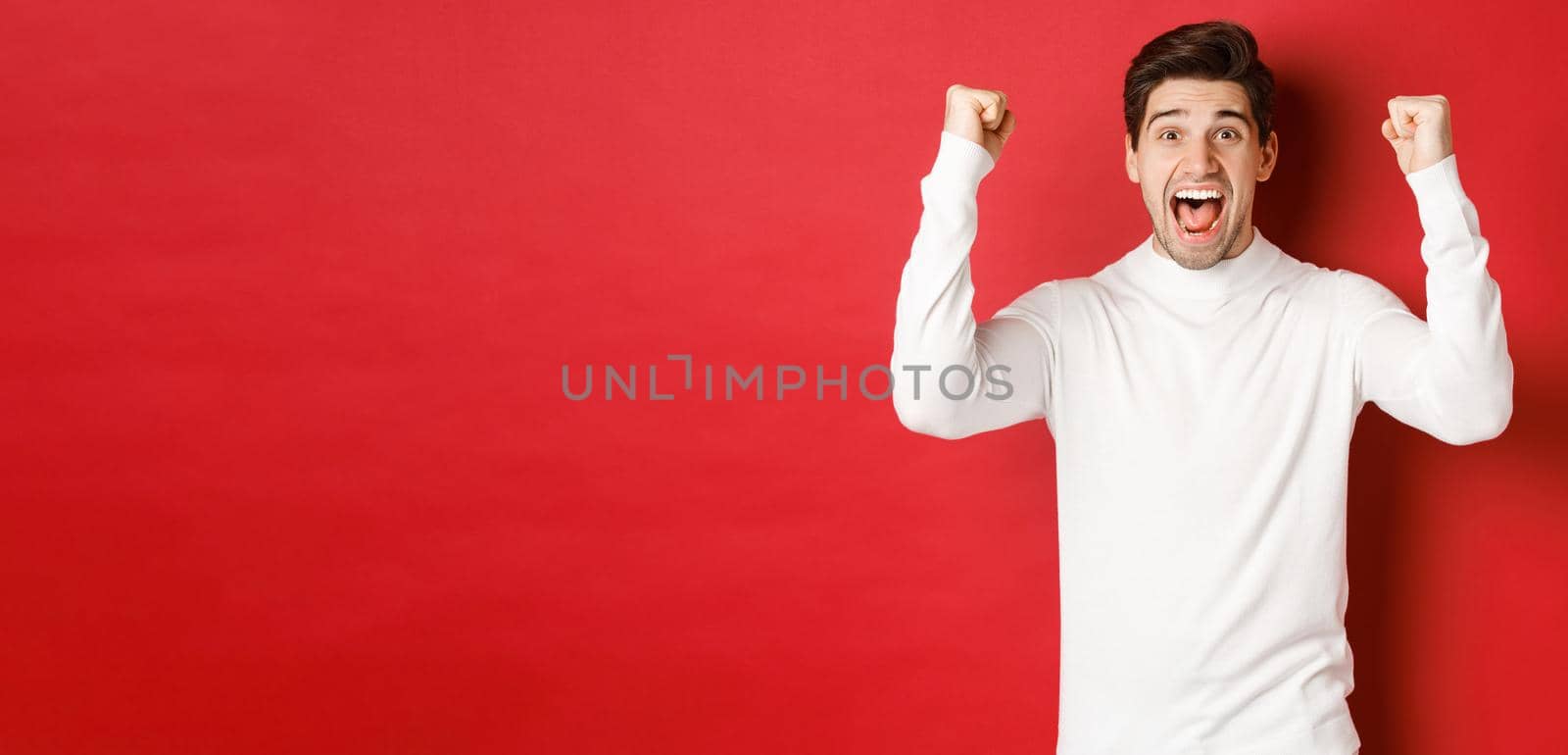 Portrait of excited lucky man in white sweater, raising hands up and triumphing, celebrating new year, standing happy against red background.