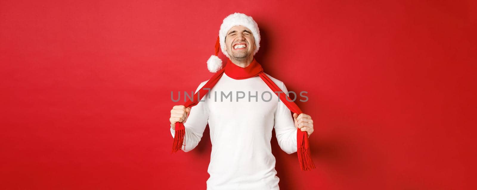Image of distressed and frustrated man in santa hat, chocking himself with scarf from sadness, standing against red background by Benzoix