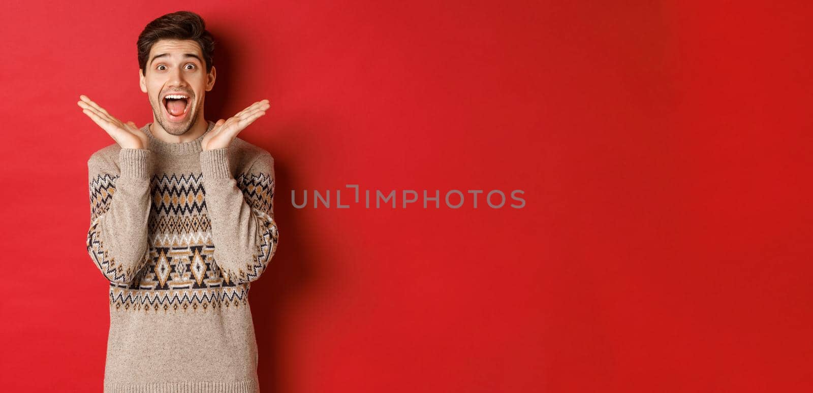 Portrait of surprised and excited guy in christmas sweater, reacting to new year present, smiling amazed, standing over red background by Benzoix