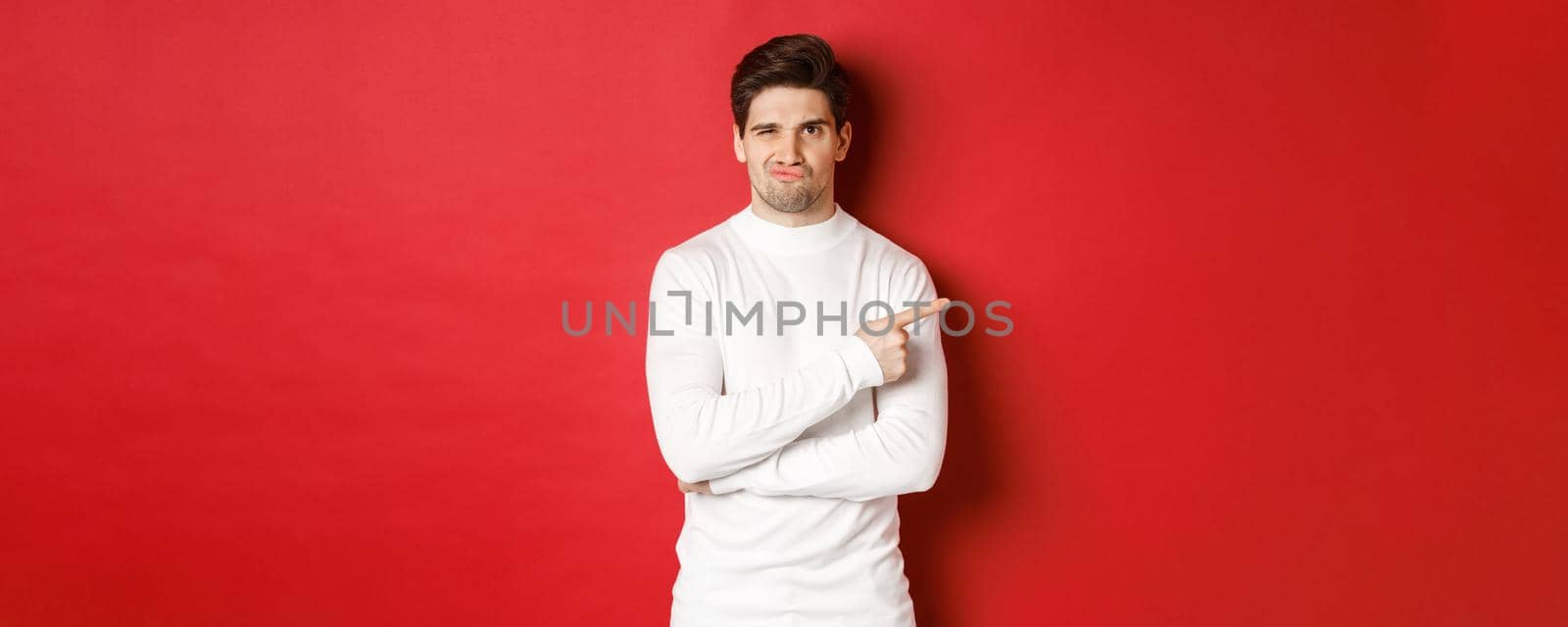Doubtful and reluctant handsome man grimacing, pointing fingers right at something bad, standing in white sweater over red background.