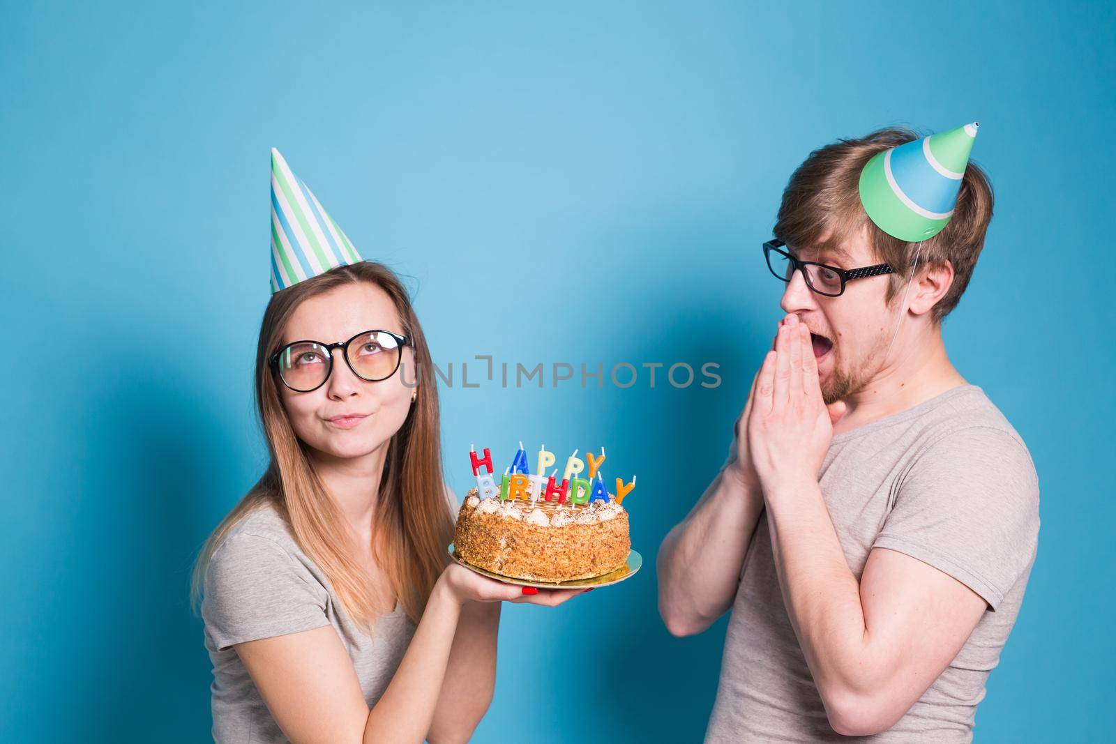 Funny young couple in paper caps and with a cake make a foolish face and wish happy birthday while standing against a blue background. Concept of congratulations and fooling around