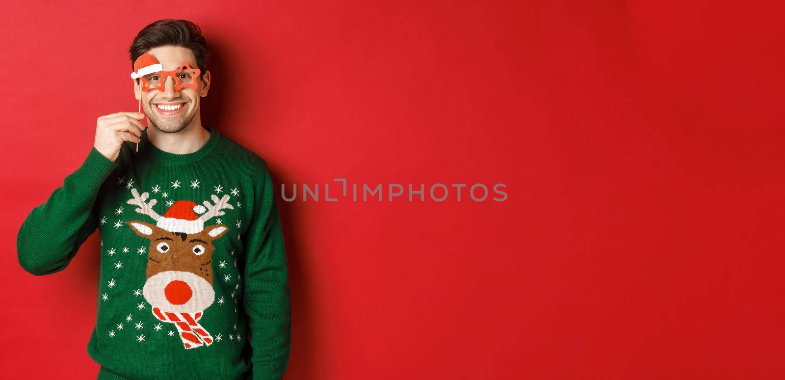 Portrait of handsome smiling man in christmas sweater and party glasses, celebrating new year and having fun, standing against red background.