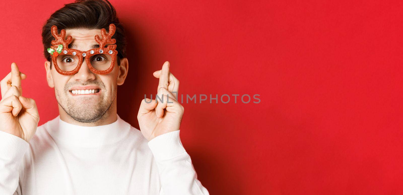 Concept of winter holidays, christmas and celebration. Close-up of nervous man in party glasses, crossing fingers for good luck and pleading, making a wish, standing over red background by Benzoix