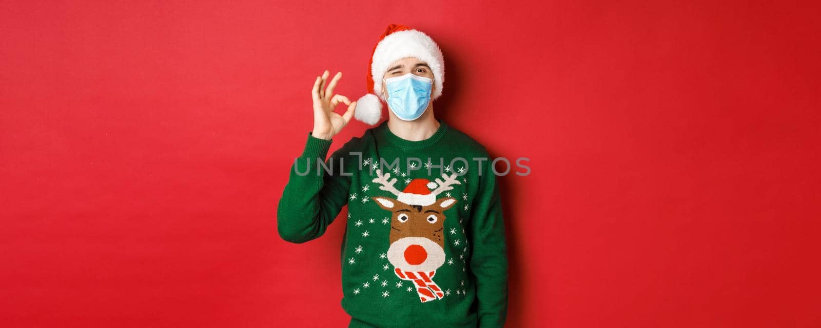 Concept of new year, covid-19 and social distancing. Cheerful man in medical mask and santa hat, showing okay sign, recommending something good, standing over red background.