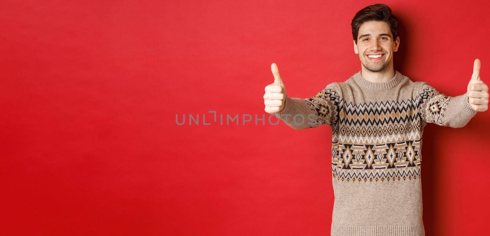 Image of handsome caucasian man in christmas sweater, showing thumbs-up in approval and smiling, wishing happy holidays, standing over red background by Benzoix