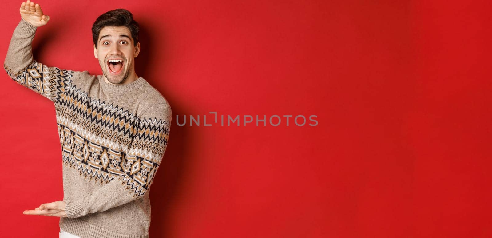 Portrait of excited and happy handsome man in christmas sweater, showing something big, holding large gift for holidays, standing over red background.