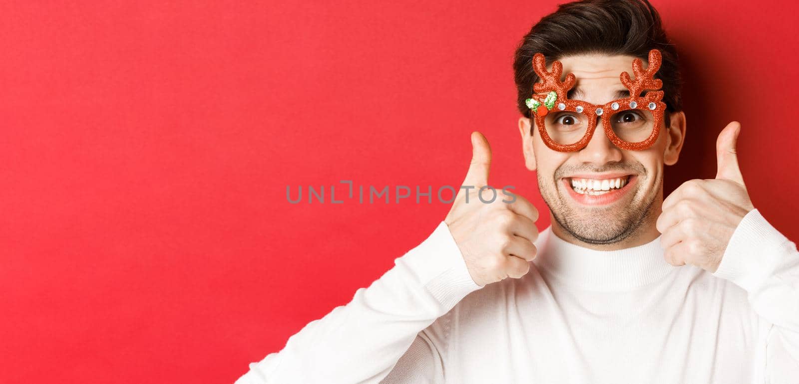Concept of winter holidays, christmas and celebration. Close-up of excited handsome man in party glasses, smiling and showing thumbs-up in approval, standing over red background by Benzoix