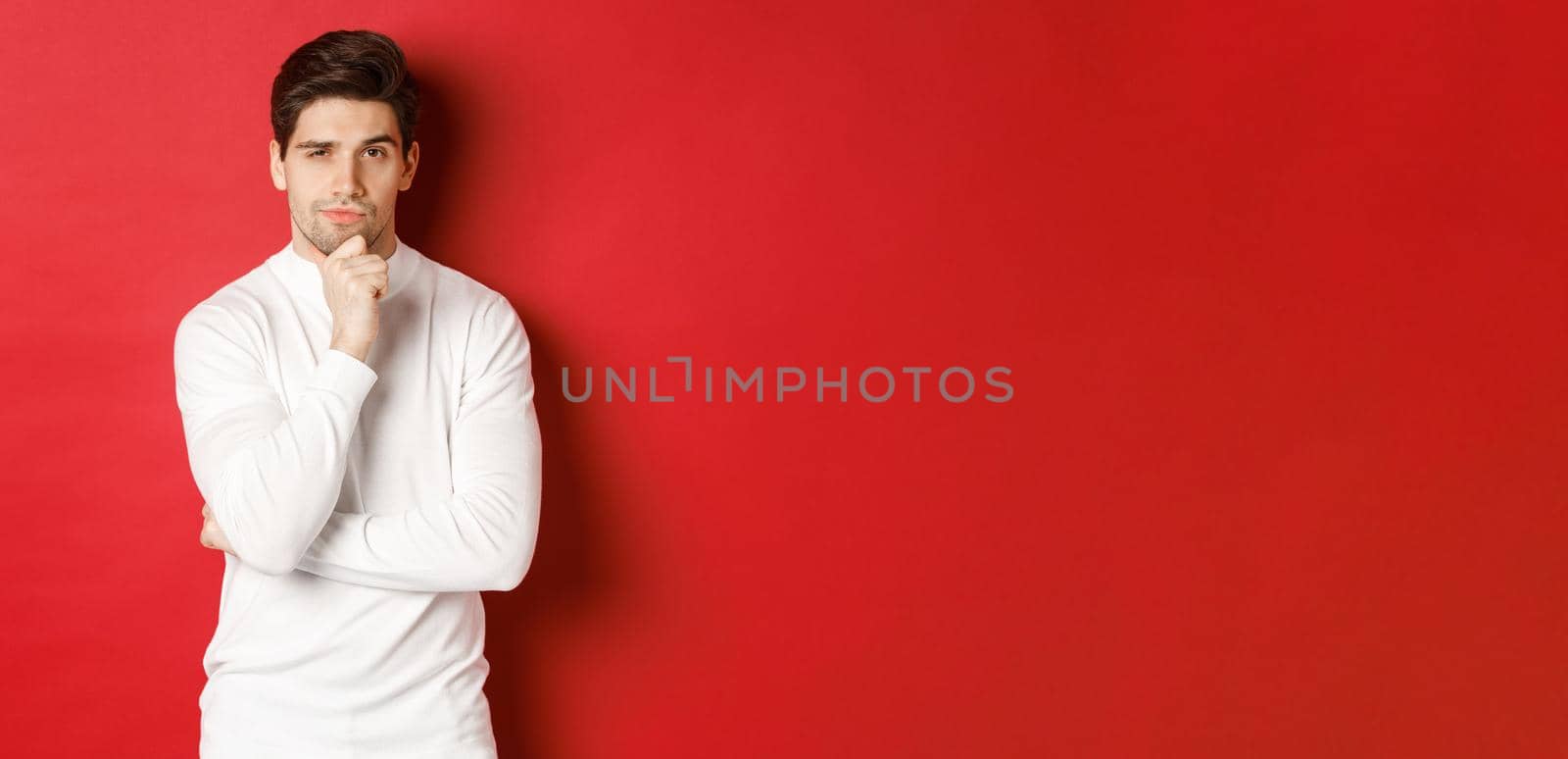Image of thoughtful handsome man making assumption, thinking and looking at camera, standing in white sweater against red background.