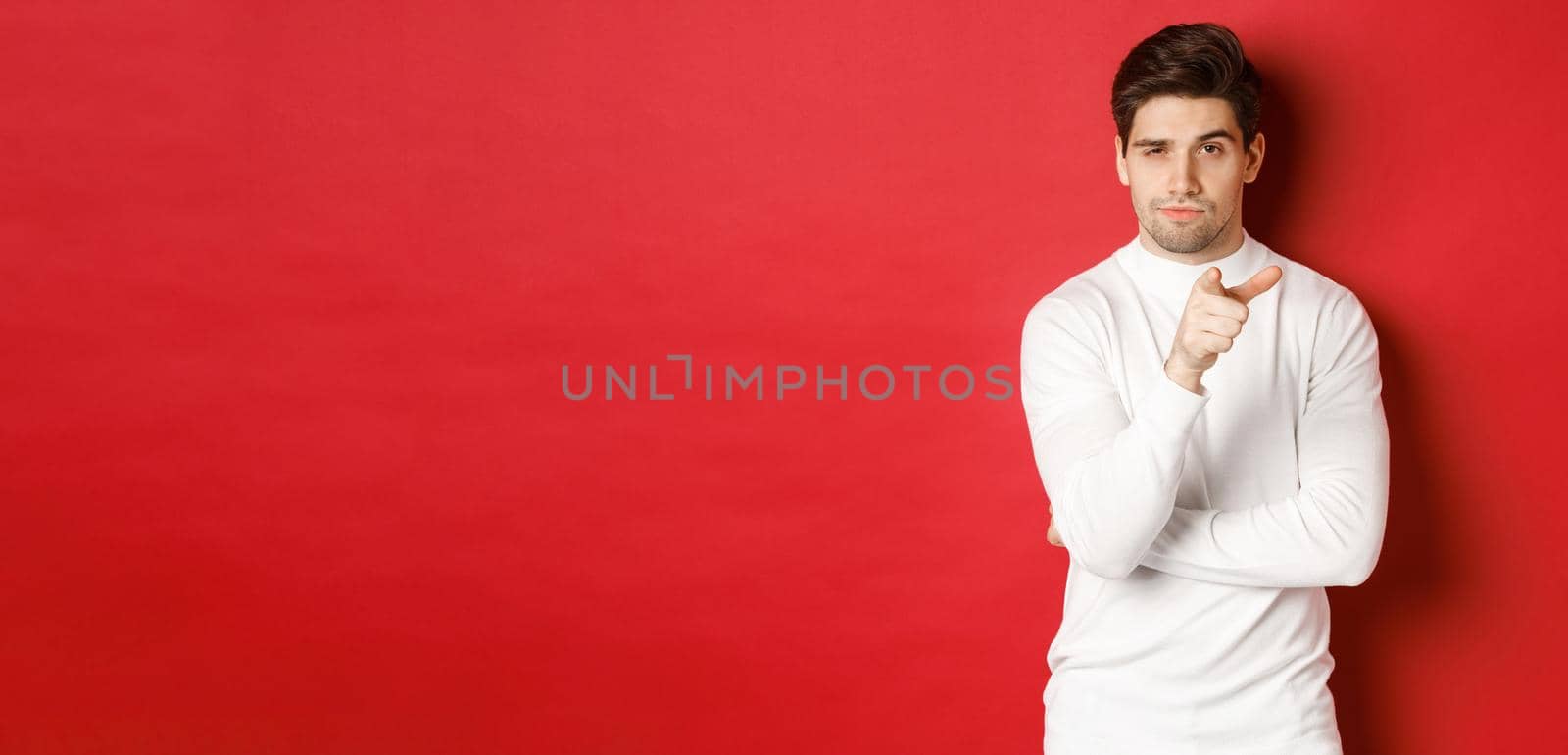 Portrait of thoughtful handsome man in white sweater, pointing at camera and making choice, standing against red background by Benzoix