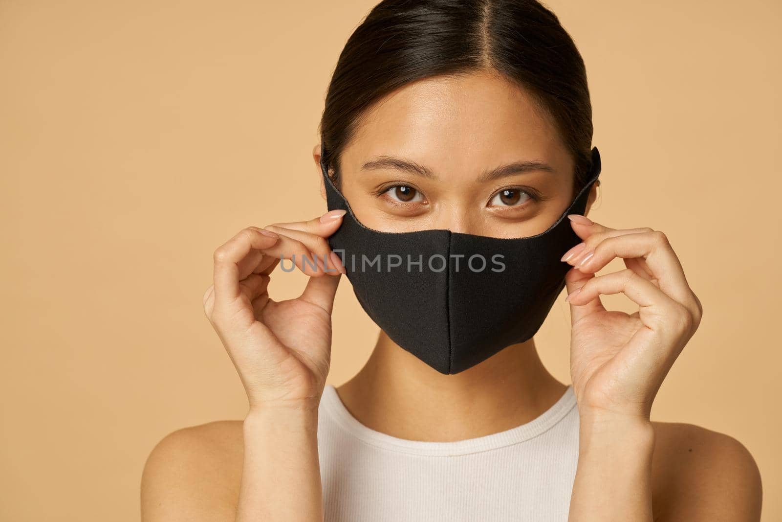 Charming young woman adjusting black facial mask and looking at camera, posing isolated over beige background. Safety, pandemic concept