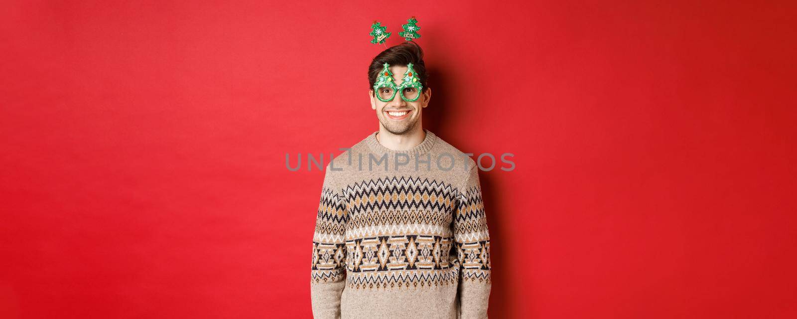 Portrait of happy and cute man in winter sweater and party glasses, celebrating new year or christmas, smiling while standing over red background.