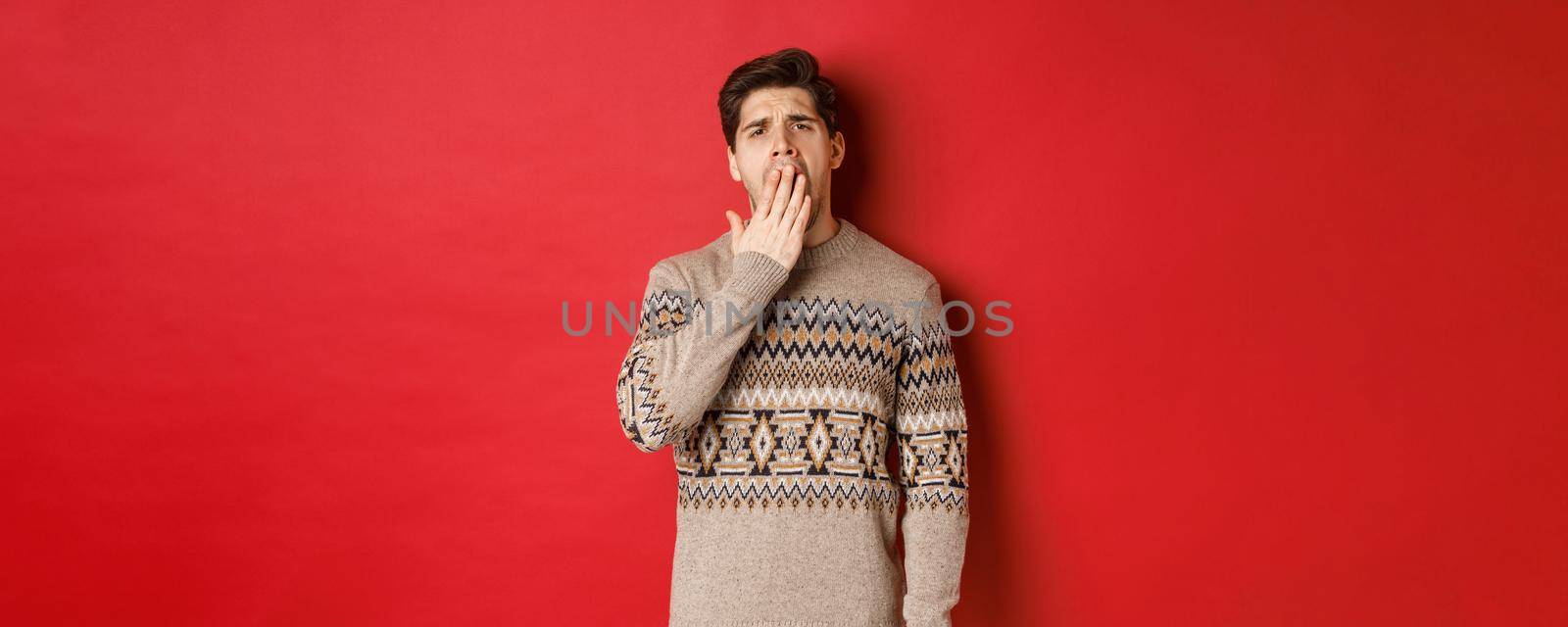 Image of tired or bored handsome man in winter sweater, yawning and covering mouth with hand, standing exhausted against red background by Benzoix