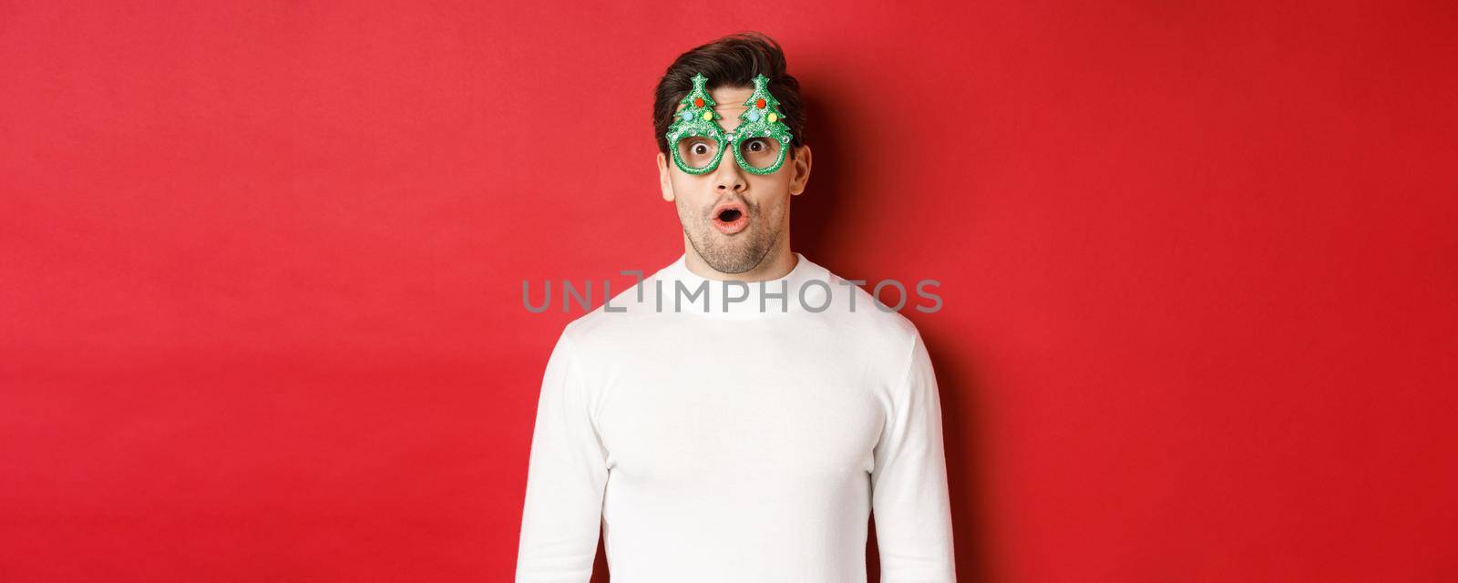 Concept of christmas, winter holidays and celebration. Close-up of surprised handsome guy in party glasses, saying wow and looking at amazing new year promo offer, red background by Benzoix