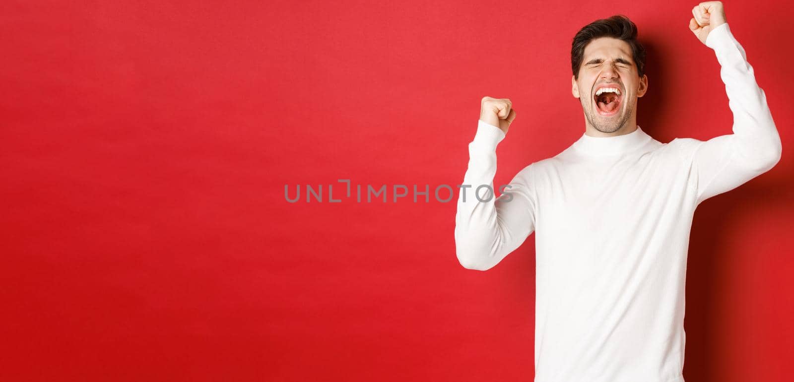 Portrait of handsome man in white sweater, feeling cheerful, celebrating victory, shouting for joy and raising hands up in victory, standing over red background by Benzoix