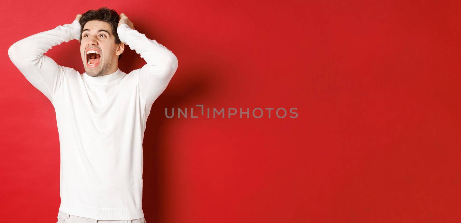 Portrait of frustrated man in white sweater, screaming in panic and looking at upper left corner, have terrible problem, standing over red background.