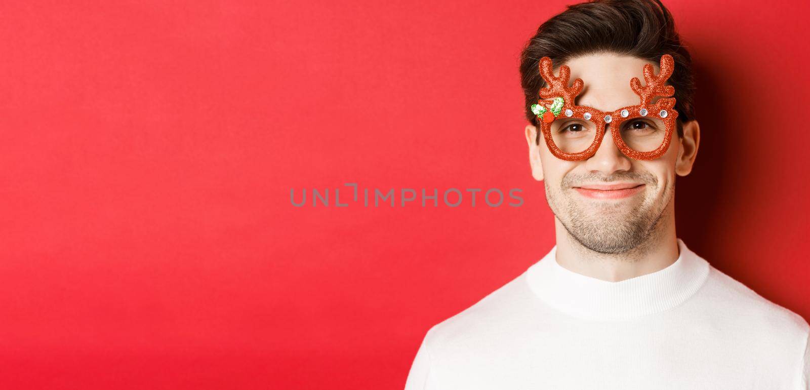 Concept of winter holidays, christmas and celebration. Close-up of attractive smiling male model in party glasses, standing against red background.