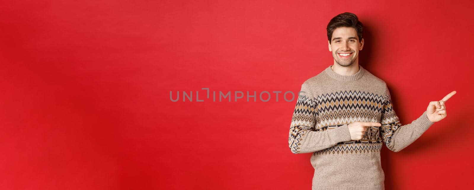 Image of attractive smiling man, wearing christmas sweater, pointing fingers right and showing new year advertisement, standing over red background by Benzoix
