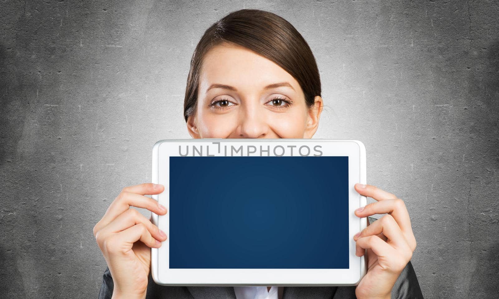 Businesswoman holding tablet computer with blank screen. Beautiful woman in business suit show tablet PC near her face. Corporate businessperson on grey wall background. Digital technology layout.