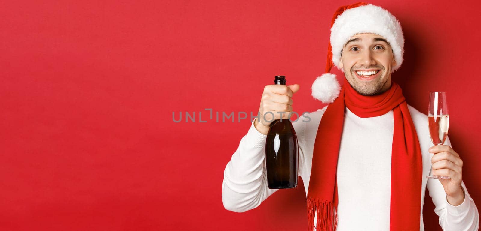 Concept of winter holidays, christmas and lifestyle. Close-up of cheerful handsome man, holding champagne bottle and glass, making toast for new year and celebrating, red background by Benzoix