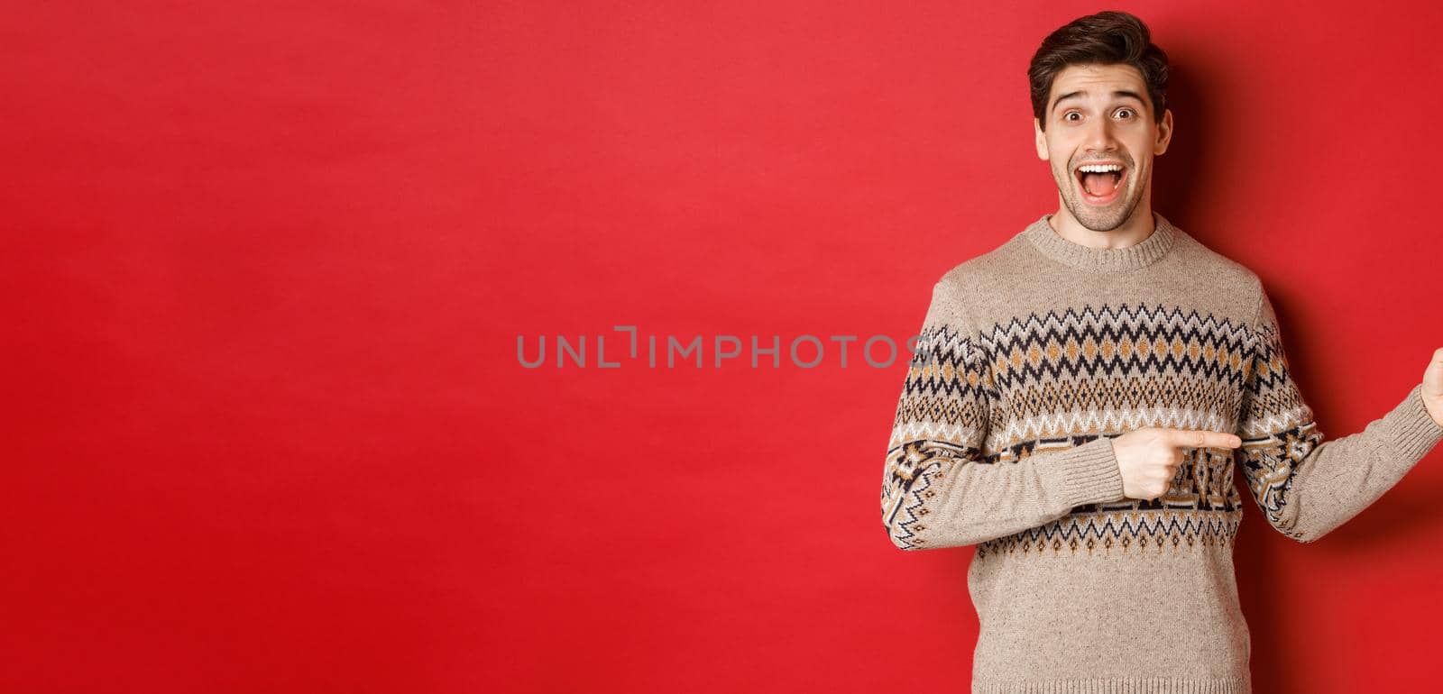 Concept of christmas celebration, winter holidays and lifestyle. Handsome man in xmas sweater pointing fingers right, smiling amazed, showing new year promo against red background by Benzoix