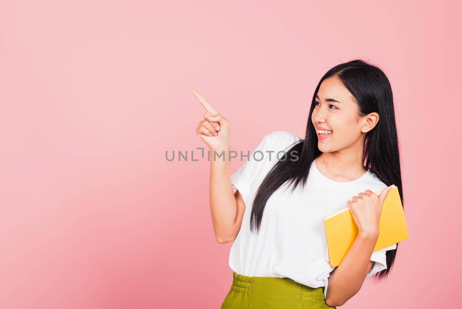 Portrait of happy Asian beautiful young woman confident smiling holding orange book open pointing finger to side copy space, studio shot isolated on pink background, education concept