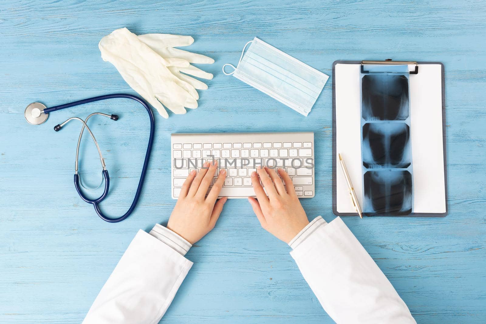 Top view of doctor hand typing at keyboard. Radiography examination in hospital. Therapist sitting at blue wooden desk with stethoscope, medical gloves and mask. Examination and consultation in clinic