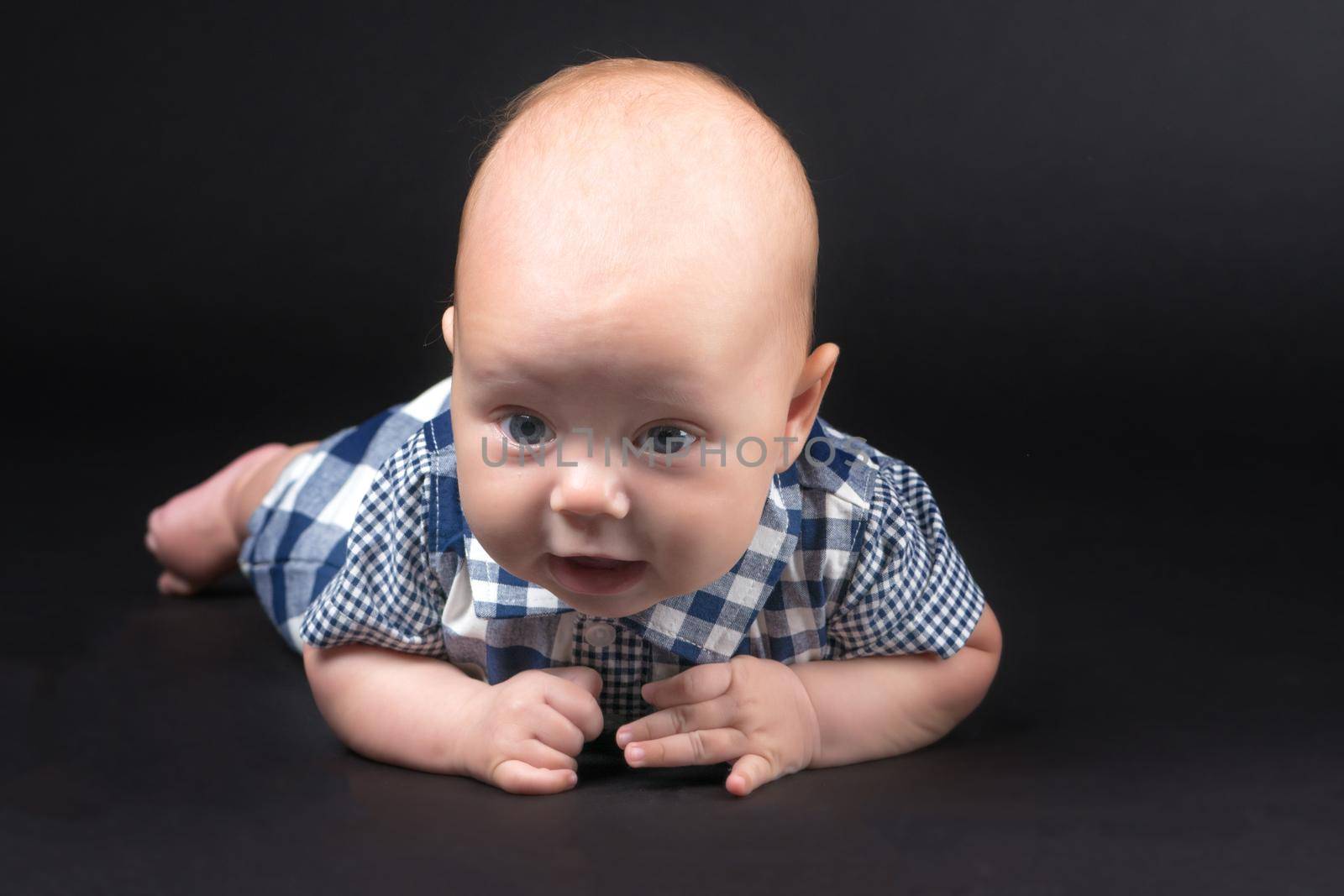 A charming baby lies on the blanket and looks into the camera on a black background. The concept of a happy childhood, the birth and upbringing of a child.