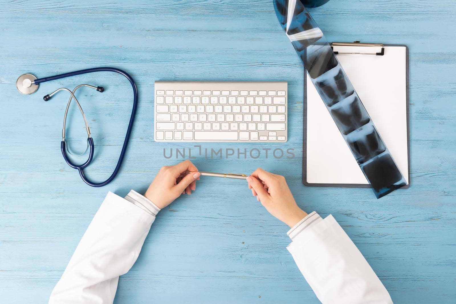 Top view of doctor hands holding pen. X-Ray radiography examination in hospital. Physician sitting at blue wooden desk. Examination and consultation in clinic. Workplace with stethoscope and keyboard.