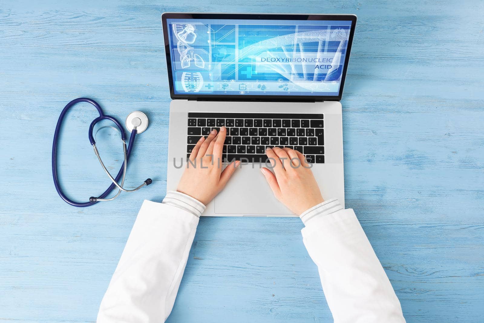 Top view of doctor hands typing at laptop computer. Therapist in white coat sitting at blue wooden desk. Online medical clinic communication with patient. Medical application and healthcare services.