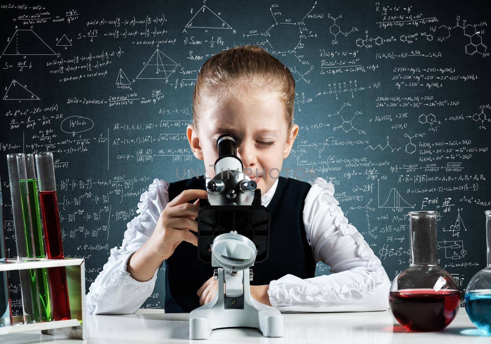 Little girl scientist looking through microscope in classroom at chemistry lesson. School chemical laboratory with glass flasks and test tubes. Schoolgirl making experiment in chemistry class.