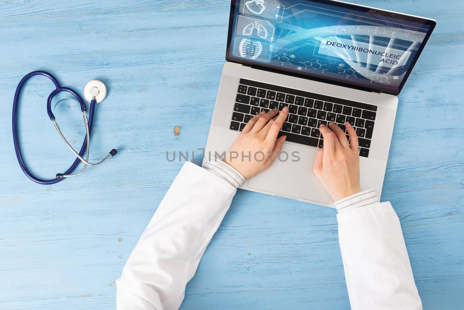 Top view of doctor hands typing at laptop computer. Therapist in white coat sitting at blue wooden desk. Online medical clinic communication with patient. Medical application and healthcare services.