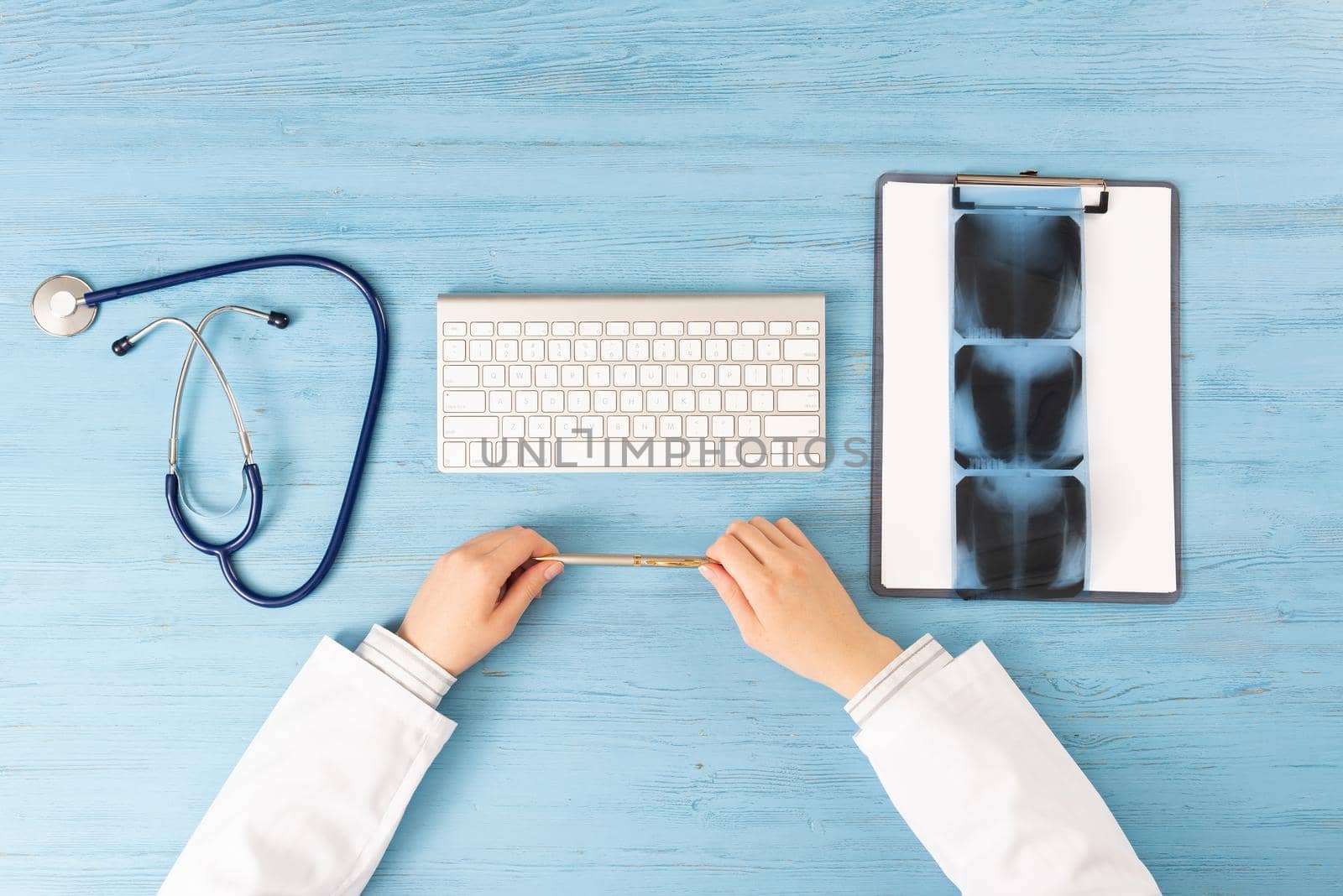 Top view of doctor hands holding pen. X-Ray radiography examination in hospital. Physician sitting at blue wooden desk. Examination and consultation in clinic. Workplace with stethoscope and keyboard.