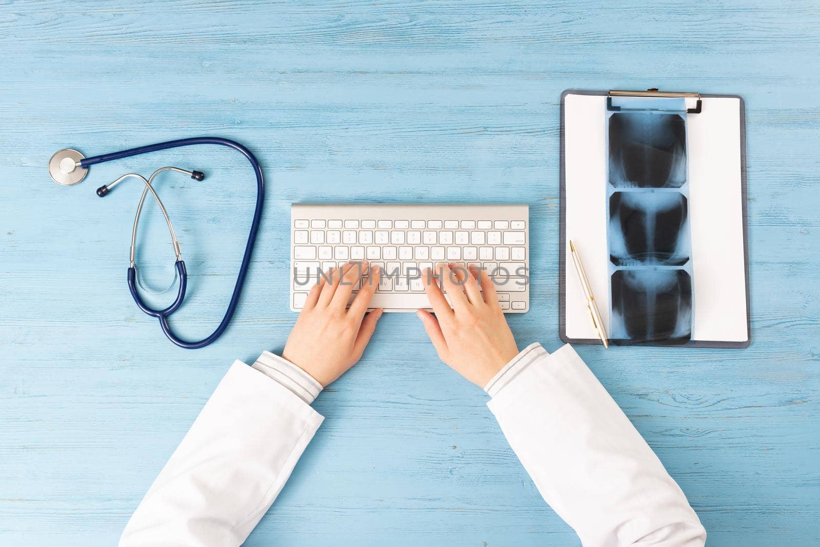 Top view of doctor hands typing at keyboard. Radiography examination in hospital. Radiologist sitting at blue wooden desk with stethoscope and x-ray scan. Examination and consultation in clinic.