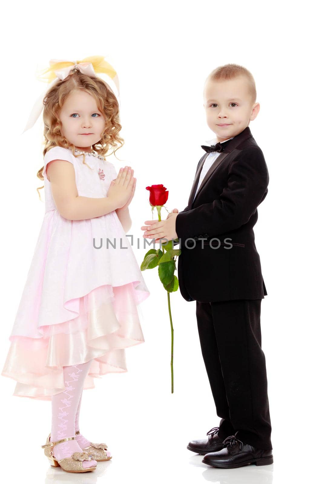 Little boy in black suit with bow tie gives a big red rose charming little girl.Isolated on white background.