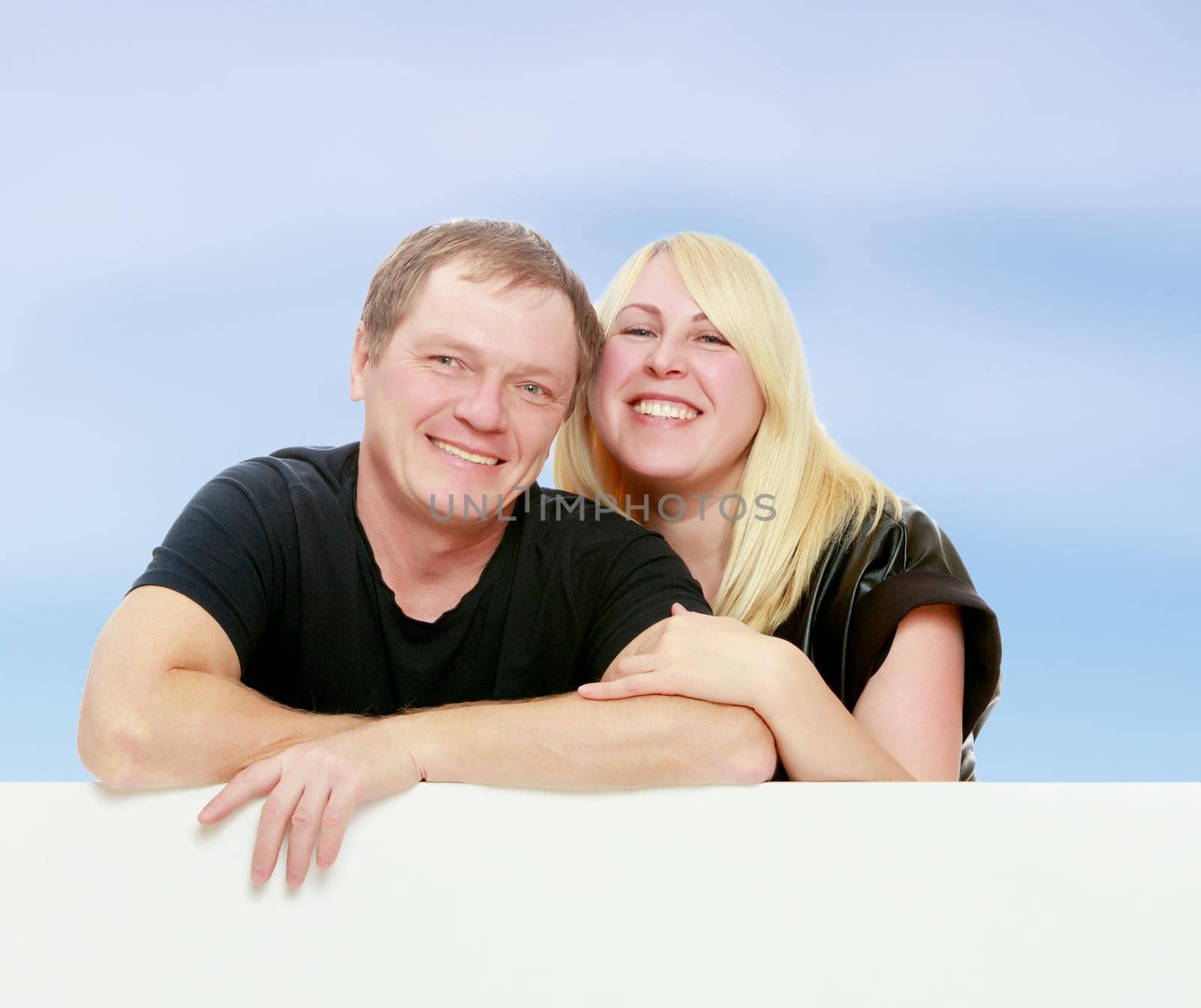 A man and a woman tenderly hugging each other. Peeping over white banner.On the background of summer blue sky and fluffy clouds.